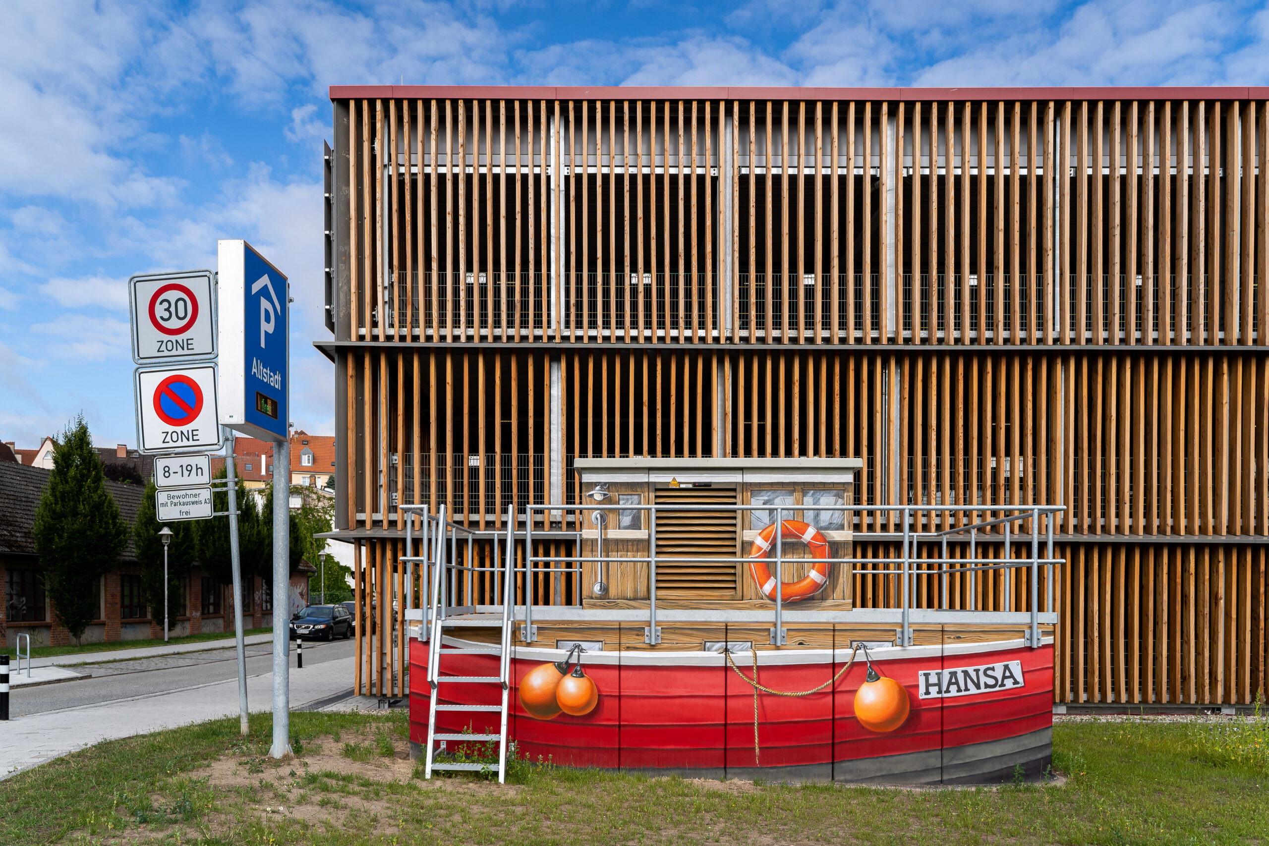 Rostocker Parkhaus Altstadt - Blick auf die Holz-Latten-Fassade aus Richtung Osten, im Vordergrund ein künstlerisch gestaltetes Trafo-Gebäude, das wie ein Fischerkahn aussieht